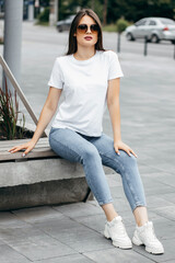 Fototapeta premium Stylish brunette girl wearing white t-shirt, blue jeans and posing sitting on a bench , urban clothing style. Street photography