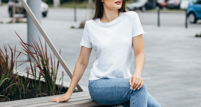 Stylish brunette girl wearing white t-shirt, blue jeans and posing sitting on a bench , urban clothing style. Street photography