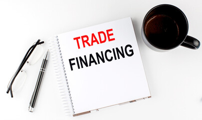 Coffee,glasses,pen and notebook written with TRADE FINANCING , on white background.