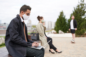 Busy young middle-eastern businessman in mask sitting on bench in city park and preparing report on laptop, coronavirus restrictions