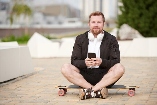 Portrait of smiling hipster guy with beard sitting on skateboard in city park and texting message on smartphone