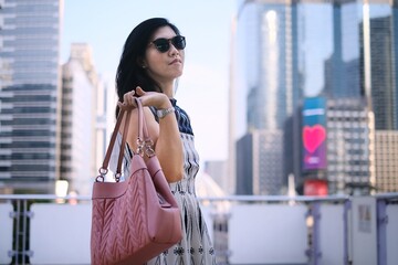 A beautiful confident Asian businesswoman in white and blue dress is holding her pink handbag, standing on an overpass walkway with the city skyline in the background.