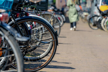 Selective focus of back wheel of bicycle in the row, Outdoor public parking area with blurred people walking in parking slot, Cycling is a common mode of transport in the Netherlands.
