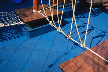 Sunlight and shadow on surface of climbing rope with vintage outdoor wooden playground equipment on blue rubber tile floor in public park area