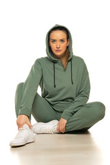Obraz premium Portrait of a young woman in sports clothes with hood, sitting and posing, against studio white background