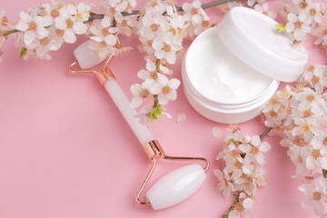 Container with bodycare and skincare cream, roller for face massage on a pink background with blooming cherry. Cosmetic facial skin care and spa. Natural treatment concept.