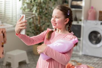 Cute little girl in pigtails holds phone takes selfie talks through video, child holds laundry liquid, rinse clothes in washing machine.