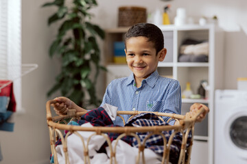 Smiling handsome little boy stands in the middle of the bathroom, laundry room, holding a large wicker basket filled with colorful clothes, son helps his mother with housework.