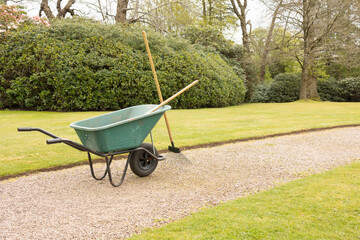 gardeners wheel barrow in green and tools on a gravel path and a well kept lawn	
