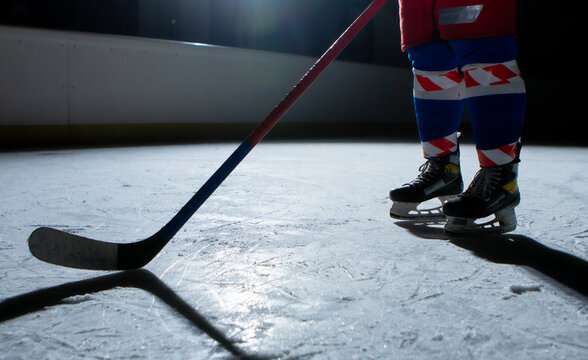 Man Hockey Player In Sports Uniform And Skates Standing On Ice Arena With Stick In His Hands. An Athlete Hones His Skills In Playing Hockey On Ice Of Stadium In Dark With Backlight. Legs Close Up.