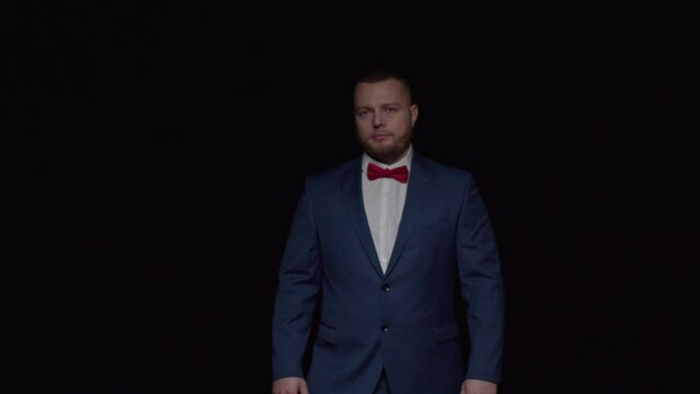 An elegant young man in a blue tuxedo, protruding from the darkness, pulls a tie and crosses his arms.