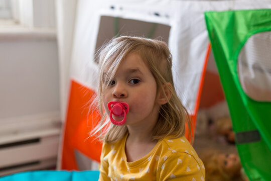 Horizontal view of adorable disheveled blond little girl playing at being a baby with a pacifier in her mouth