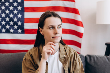 Portrait of smiling american young brunette woman sitting on comfy couch at home on background USA flag. Calm free gen z hipster girl 20s old year celebrating freedom on Independence Day concept