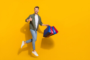 Fototapeta na wymiar Full length body size view of handsome trendy cheery guy jumping buying gifts showing thumbup isolated over bright yellow color background