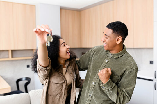 Multiracial woman holding home keys while hugging boyfriend in new apartment. Lovely couple feeling ecstatic about their recent purchase of new home - Powered by Adobe