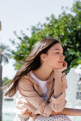 dreamy and positive brunette woman holding hand near face while sitting outdoors.