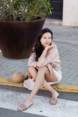 brunette woman sitting on pavement near straw hat and huge flowerpot.