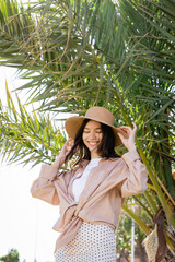 cheerful brunette woman adjusting straw hat under palm tree.