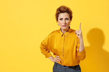 Elderly strict woman point index finger up, warns, notifies, recommends something. Photo of attractive woman in yellow shirt on yellow background