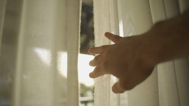 Hand slowly opens window curtain as sun light shines through into room or house. Arm enters frame to move shade or curtain as light flares camera. Warm sunny day. Hope and faith symbol positive vibe