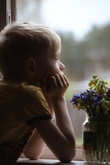 A blond boy sits by the window, leaning on the windowsill. A bouquet of spring flowers stands in a jar on the windowsill.