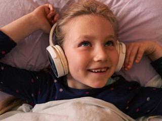 Blonde child girl in headphones learning language listening to music podcast with smartphone online...
