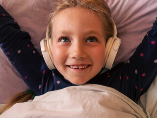 Blonde child girl in headphones learning language listening to music podcast with smartphone online in bed at home. Cute kid listens audio book tales. Distance elementary school lesson internet gadget