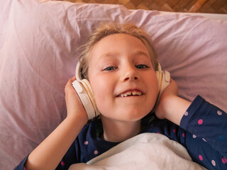 Blonde child girl in headphones learning language listening to music podcast with smartphone online...