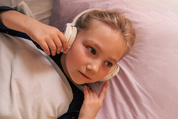 Blonde child girl in headphones learning language listening to music podcast with smartphone online...