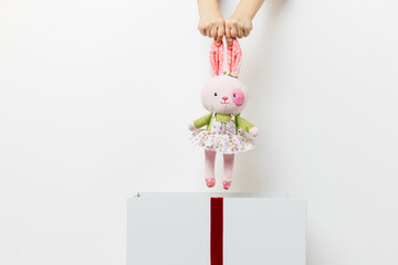 beautiful children's hands put a toy pink bunny in a white box with a red ribbon on an isolated white background. concept of gift, surprise