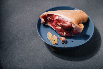 Raw pork knuckle on blue plate, seasonings, garlic, pepper set, bay leaf on grey background, close up. Cooking of meat dish, meal and flavourings, cuisine.