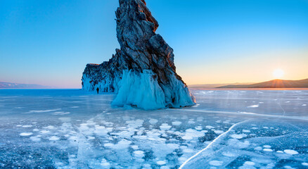 A granite rock with steep slopes rises above a frozen lake with reflection on the ice at sunset - ...