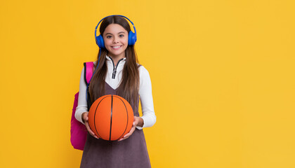 cheerful kid in headphones with school backpack and basketball ball on yellow background