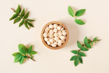 Fototapeta na wymiar Directly Above Shot Of Pills and green leaves. Tablets in wooden bowl on beige neutral colors background. Homeopathic medicine. Organic medical capsules with herbal plant. Table top view. Copy space