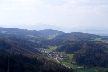 Fototapeta premium Panoramic view from local mountain Uetliberg with valley, village, agricultural fields and Swiss Alps in the background on a blue cloudy spring day. Photo taken April 14th, 2022, Zurich, Switzerland.