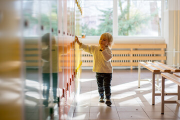 Sweet blonde toddler boy standing in front of a lockers in kindergarden or school hallway