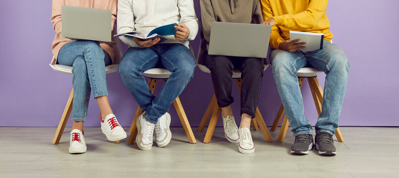 Student lifestyle. Young people with laptops and notebooks sitting in row on chairs on background of purple wall. Legs of students in casual clothes who use manuals and gadgets to prepare for exam.