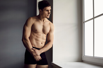 Portrait of handsome young man with sporty body posing in underwear standing by window. Caucasian...