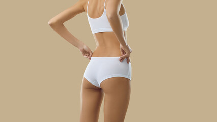 Rear view. Cropped image of female fit body in white cotton underwear isolated on beige background. Back view of unrecognizable slender skinny woman posing in top and panties with high waist. Banner.