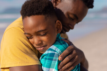 Close-up of african american boy with eyes closed embracing young father at beach on sunny day