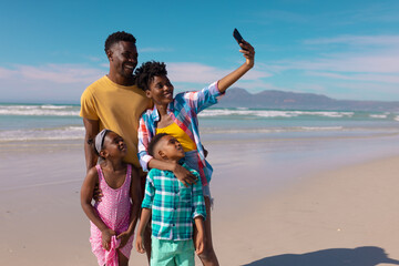 Happy african american young woman taking selfie with man, son, daughter over smartphone at beach
