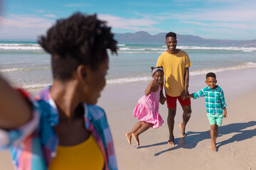 African american young woman looking at happy son, daughter and boyfriend at beach against sky