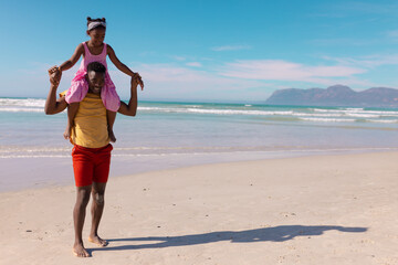 African american young man carrying daughter on shoulders while walking at beach against blue sky