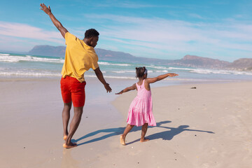 Rear view of playful african american young man and daughter with arms outstretched running at beach - Powered by Adobe