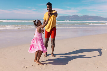 Happy african american young father dancing with daughter at beach against sea and sky on sunny day