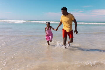 Playful african american young father and daughter holding hands and running in sea against sky