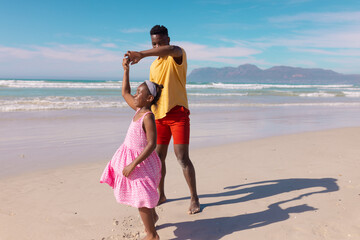 African american young man dancing with happy daughter at beach against sea and sky on sunny day