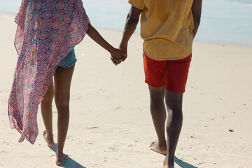 Rear view low section of african american young couple holding hands while walking on sand at beach - Powered by Adobe