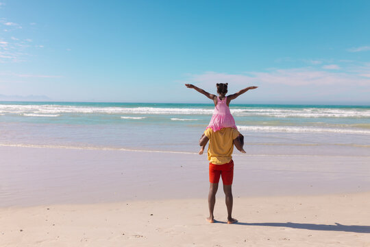 Rear view of african american girl with arms outstretched sitting on father's shoulders at beach