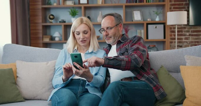 Mature couple at home using smart phone together and smiling. Man and woman relaxing on sofa and shopping on internet, watching funny videos, use social media.